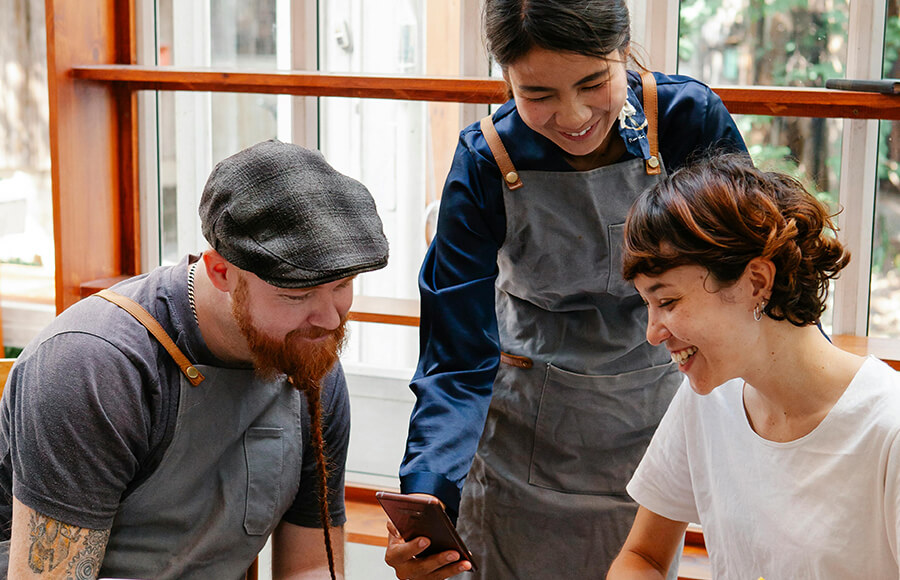 Three people in aprons huddled together, examining a cell phone screen with happy expressions.
